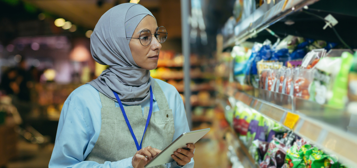 Woman Seller In Super Market In Hijab With Tablet Checking Products Using Pocket Computer, Muslim Woman Near Shelves With Products And Goods