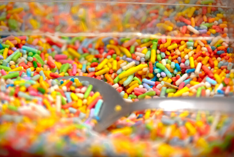 Colorful sweet chocolate sprinkles in a clear container with a spoon inside.